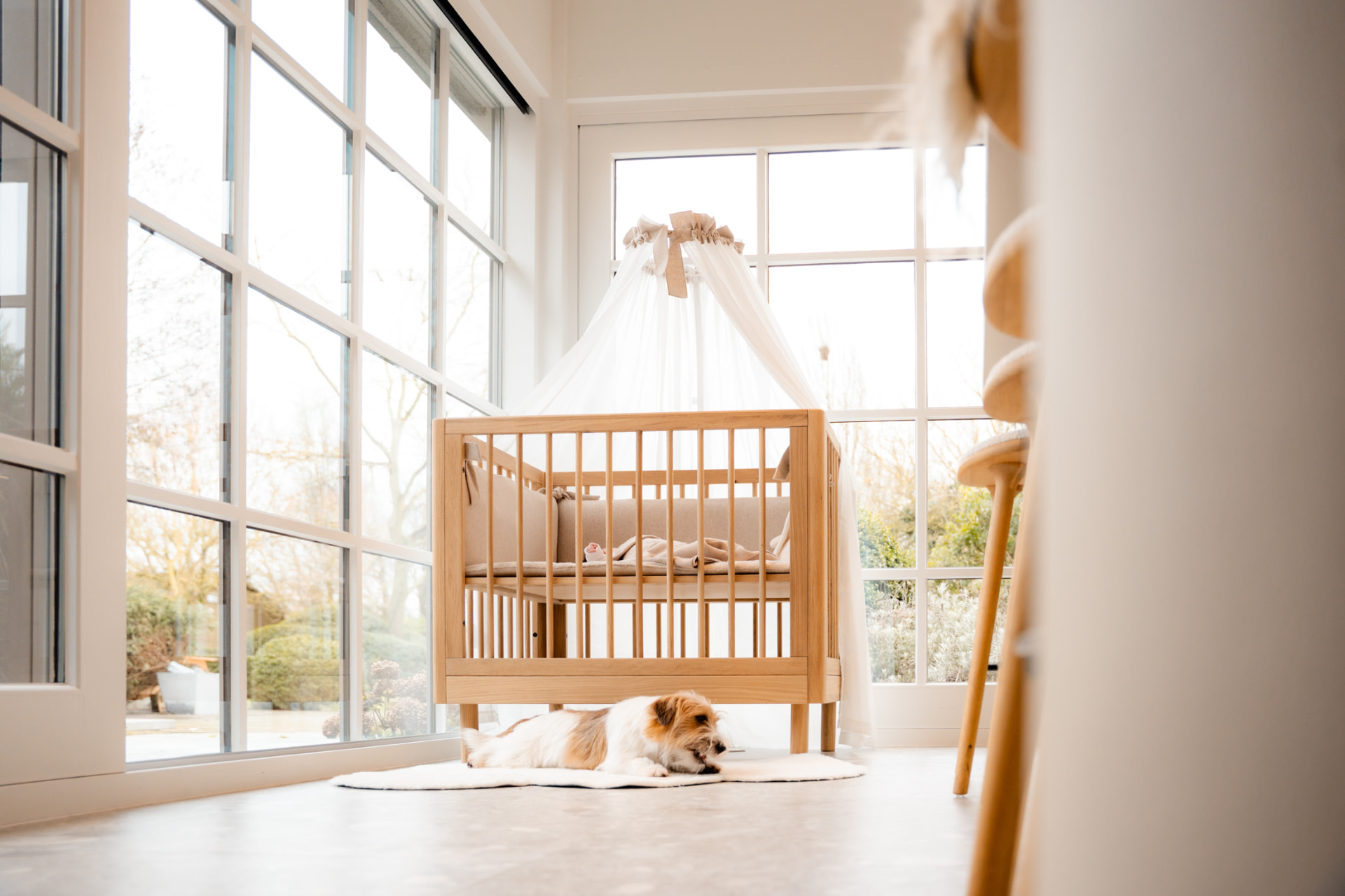 Houten babybed met hemel bij grote ramen in een lichte kinderkamer